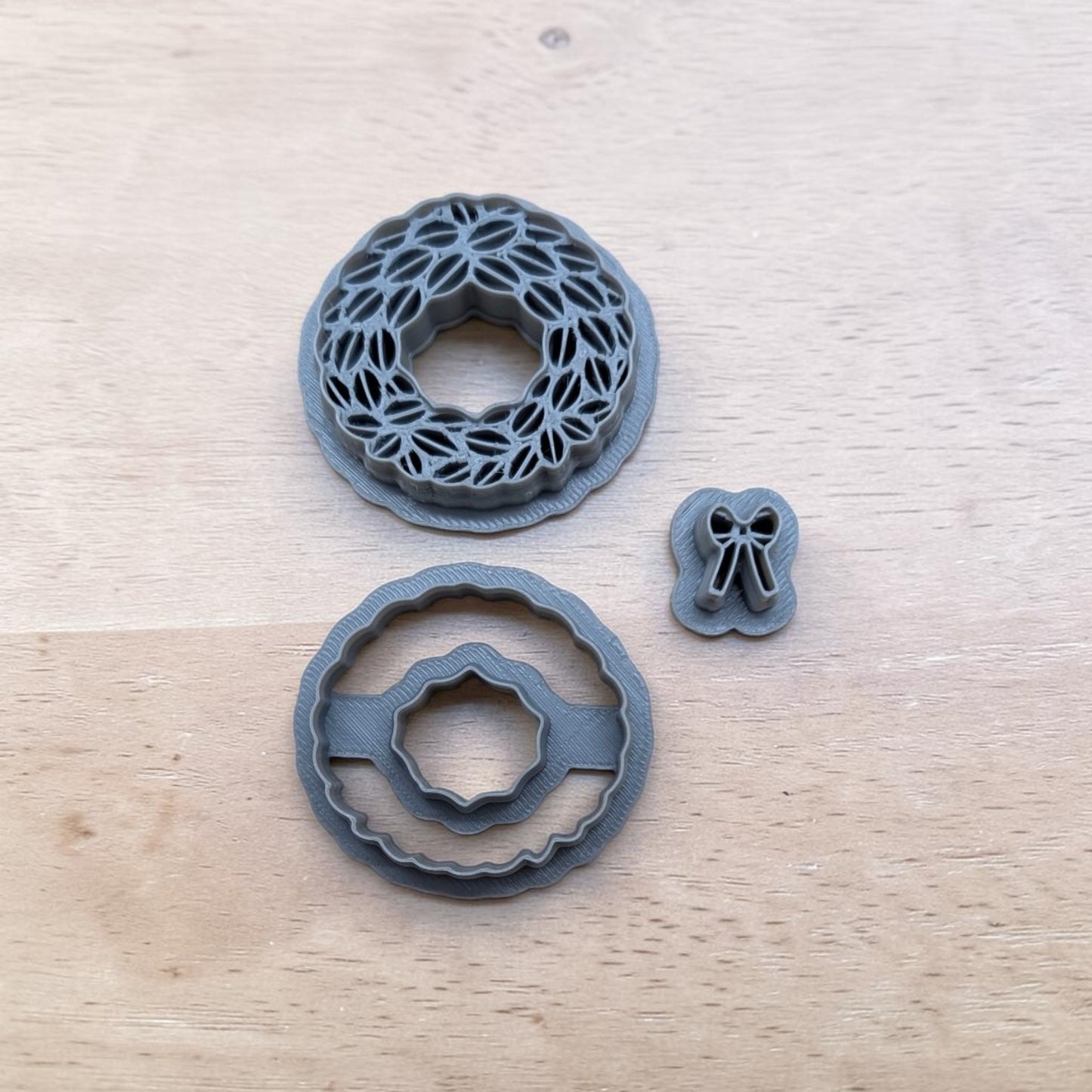 Three gray plastic cookie cutters, including a floral wreath design, a scalloped circular cutter, and a small bow cutter, arranged on a light wood surface.