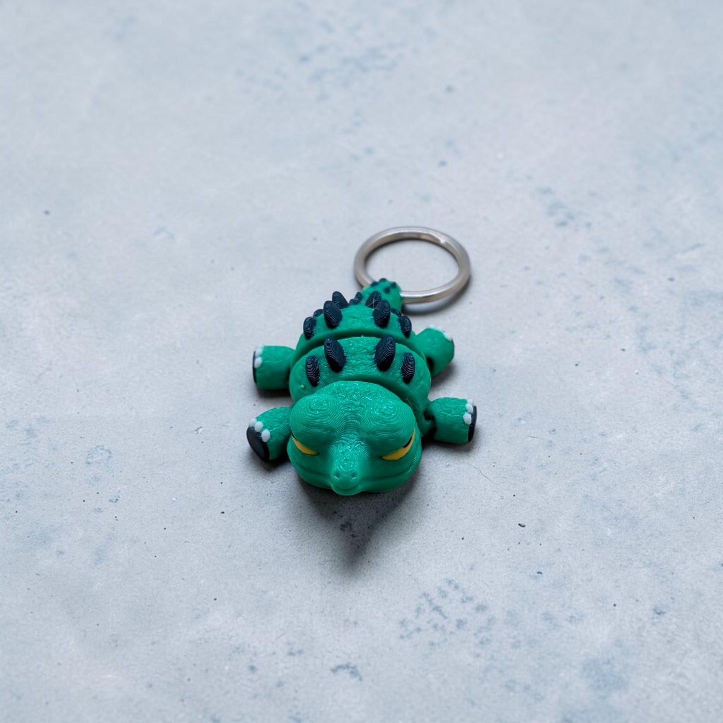 Mini Flexi Alligator keychain add‑on featuring green articulated reptile with navy spikes and yellow eyes, handmade desk buddy toy on gray surface.