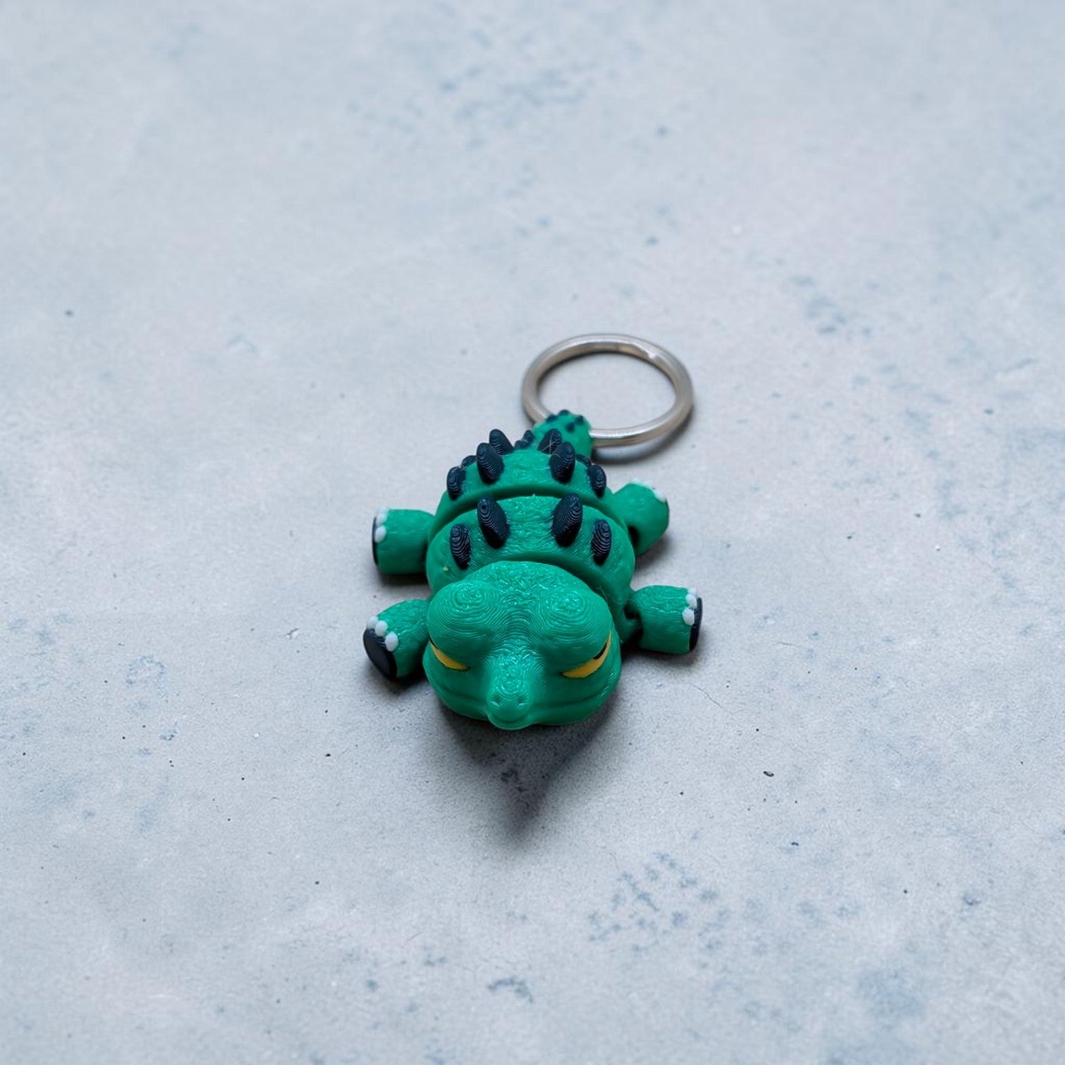 Mini Flexi Alligator keychain add‑on featuring green articulated reptile with navy spikes and yellow eyes, handmade desk buddy toy on gray surface.
