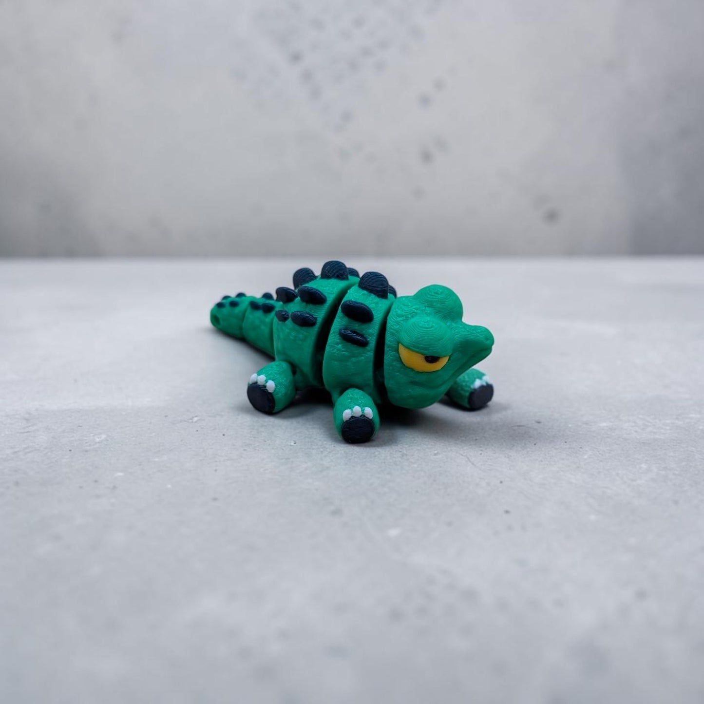 Close‑up of Mini Flexi Alligator articulated desk buddy toy showing segmented tail, navy spikes, and yellow eyes, handmade polymer clay reptile figurine.