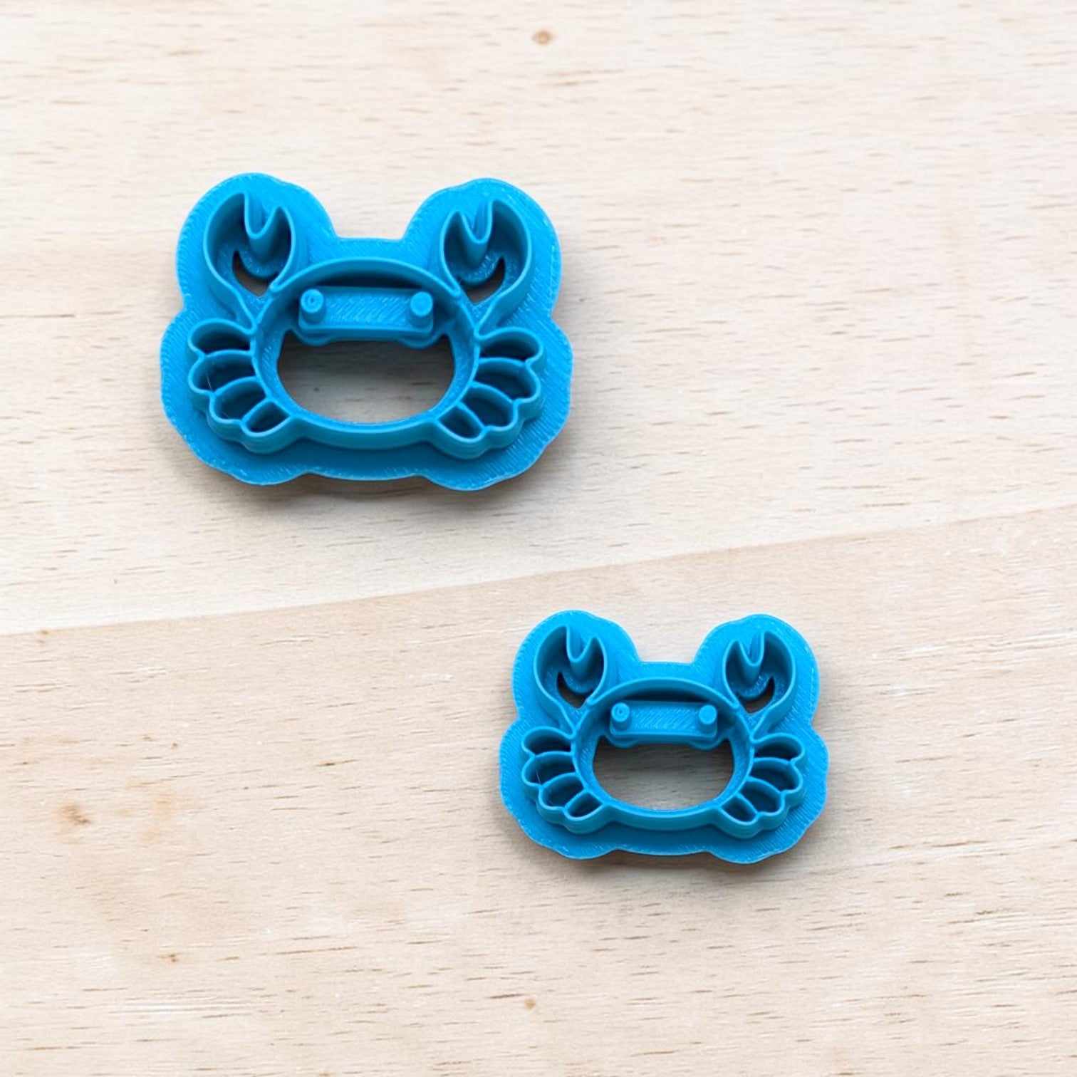 Two blue cookie cutters shaped like crabs on a light wooden surface.