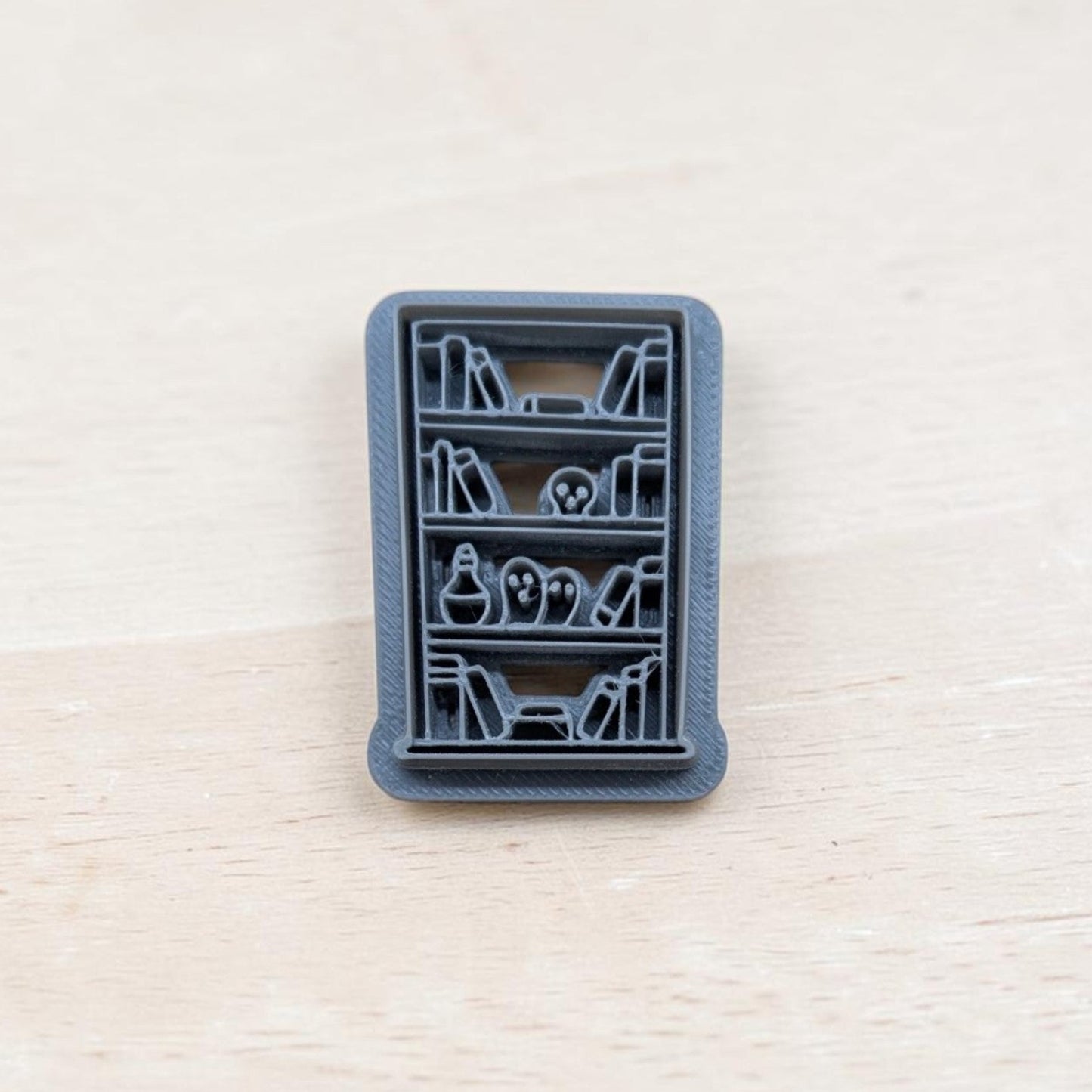 Haunted bookshelf cookie cutter – 3D printed polymer clay tool featuring spooky books, skull, and potion bottle design on shelves.