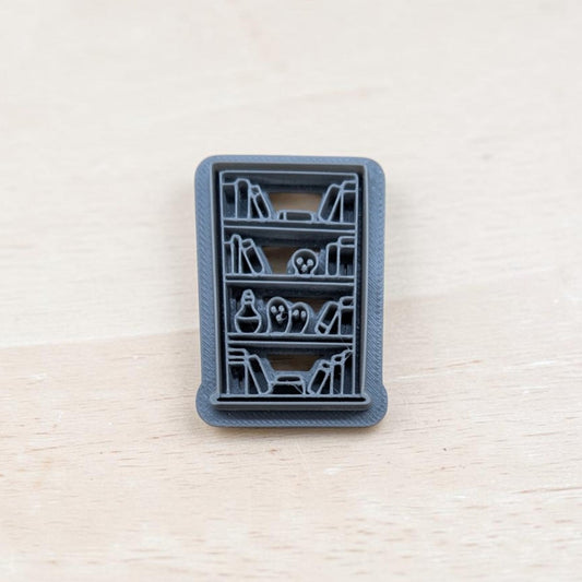 Haunted bookshelf cookie cutter – 3D printed polymer clay tool featuring spooky books, skull, and potion bottle design on shelves.