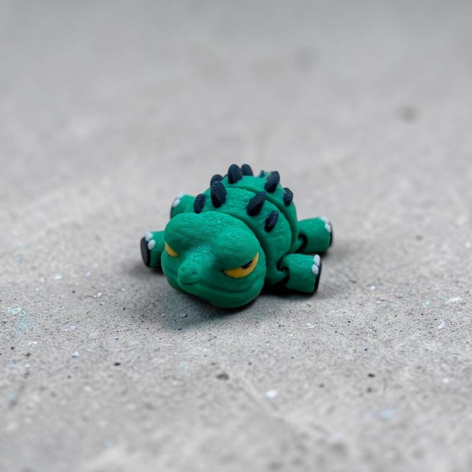 Single Mini Flexi Alligator articulated desk buddy toy in green with navy spikes and yellow eyes, handmade reptile figurine on textured gray background.