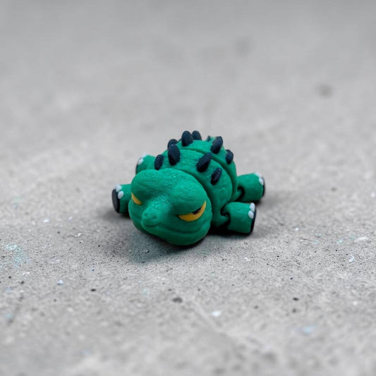 Single Mini Flexi Alligator articulated desk buddy toy in green with navy spikes and yellow eyes, handmade reptile figurine on textured gray background.