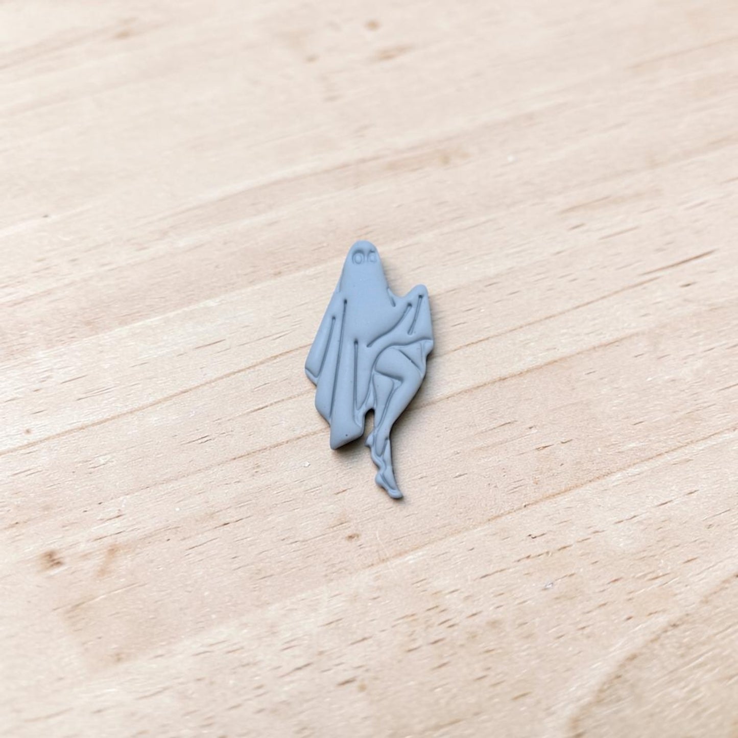 A small light blue-gray polymer clay charm shaped like a sheet ghost, placed on a light wood surface.